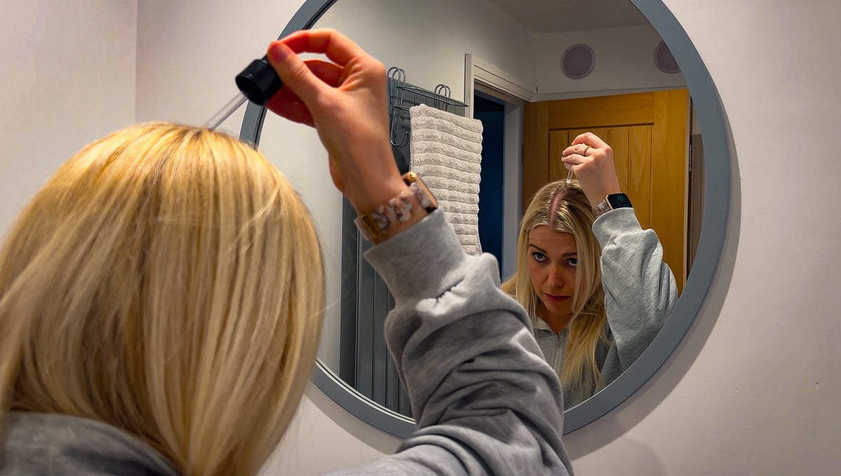 Woman applying hair growth serum to thinning hair while checking scalp in mirror