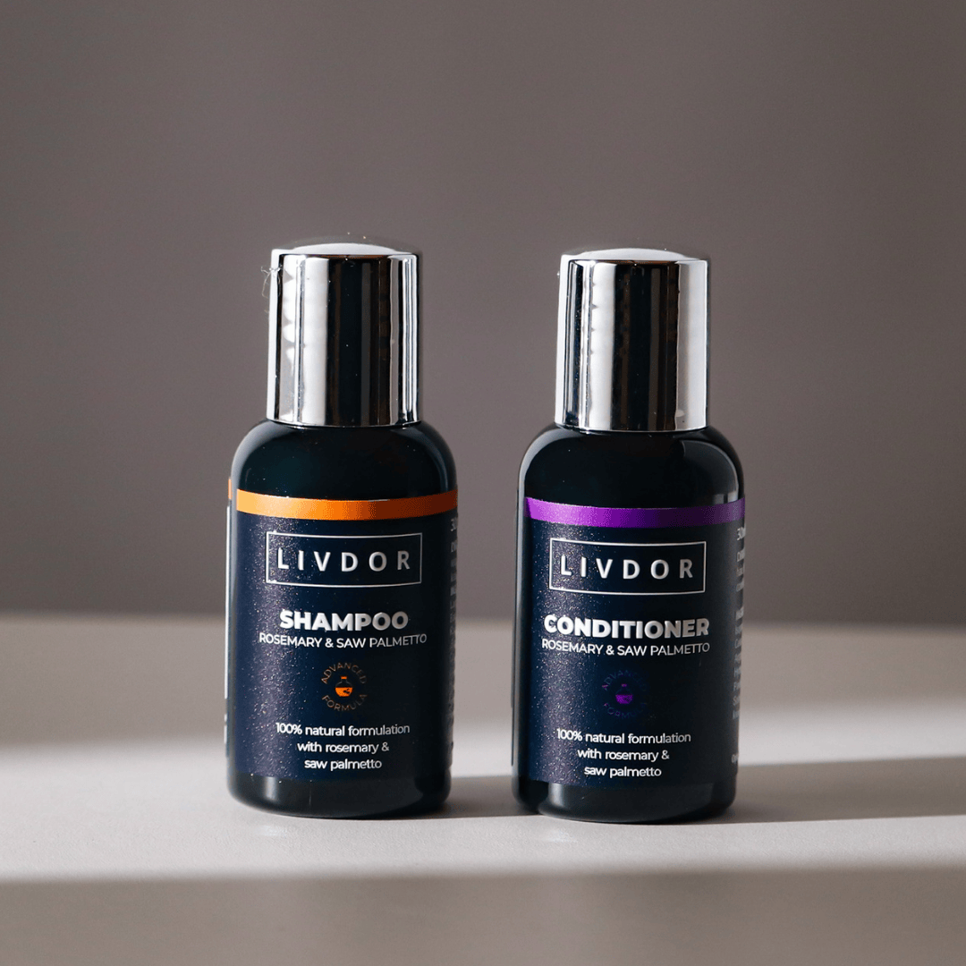 Two dark bottles of travel size LIVDOR haircare products, specifically Shampoo and Conditioner, with metallic silver caps, standing side-by-side on a light surface against a neutral gray background.